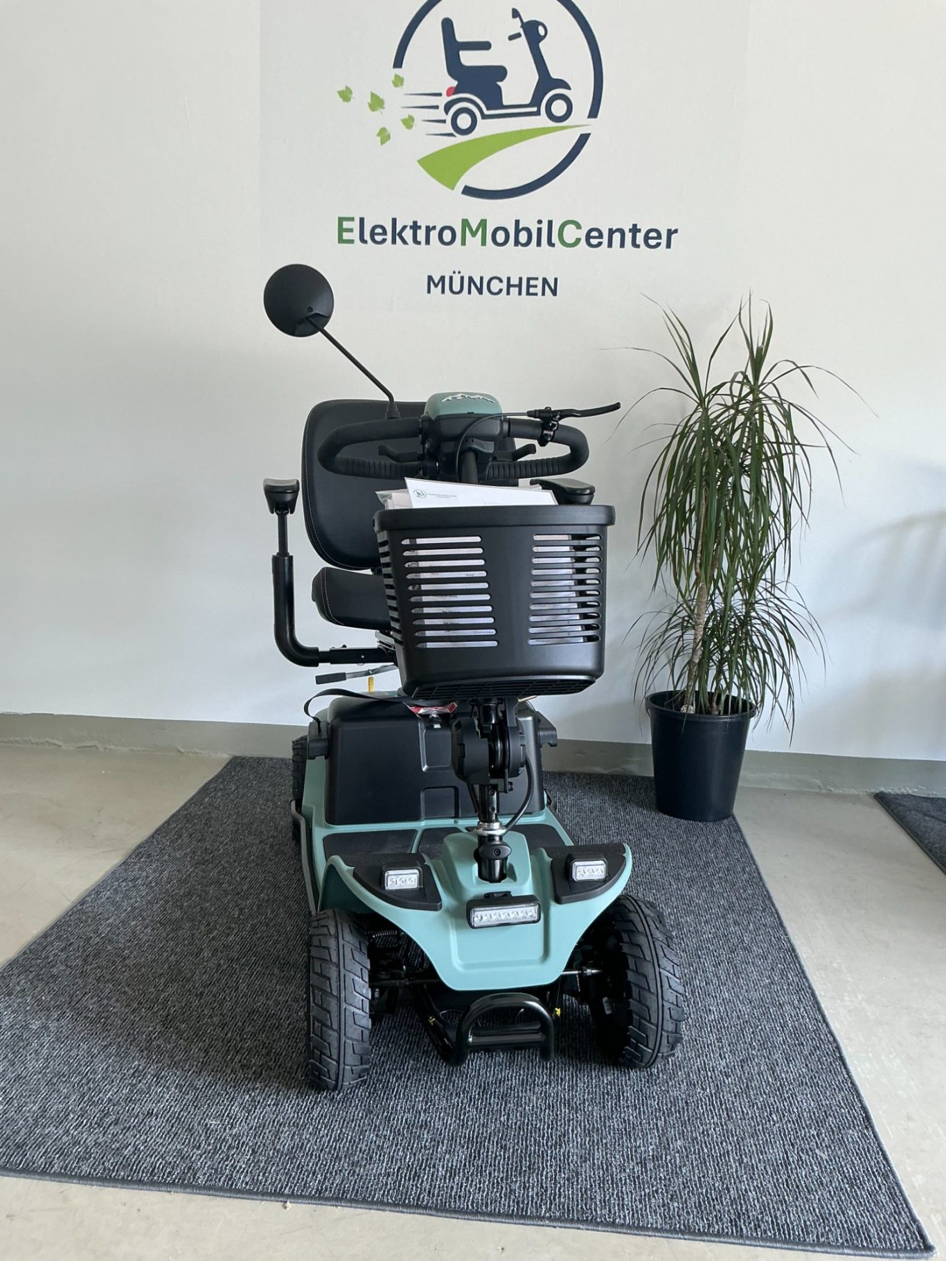 Elektrischer Rollstuhl mit Korb, auf einem Teppich vor einer Wand mit Logo.