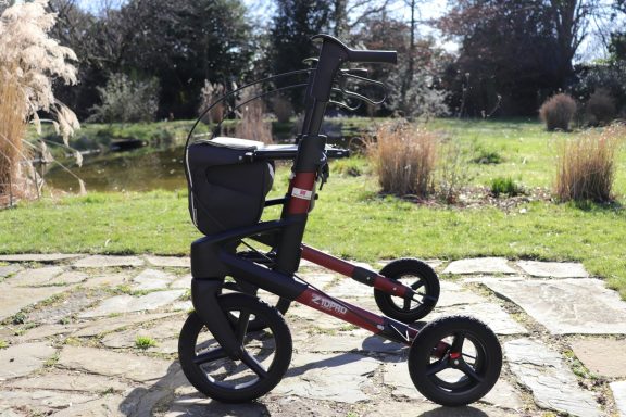 Rollator mit drei Rädern und einem Sitz, im Freien vor einer grünen Landschaft.