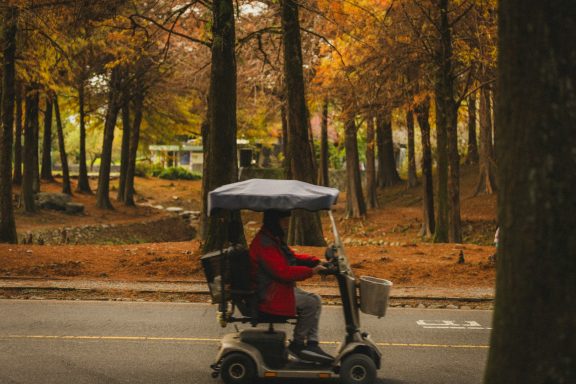 Person in rotem Mantel fährt mit Elektrorollstuhl durch einen herbstlichen Park.