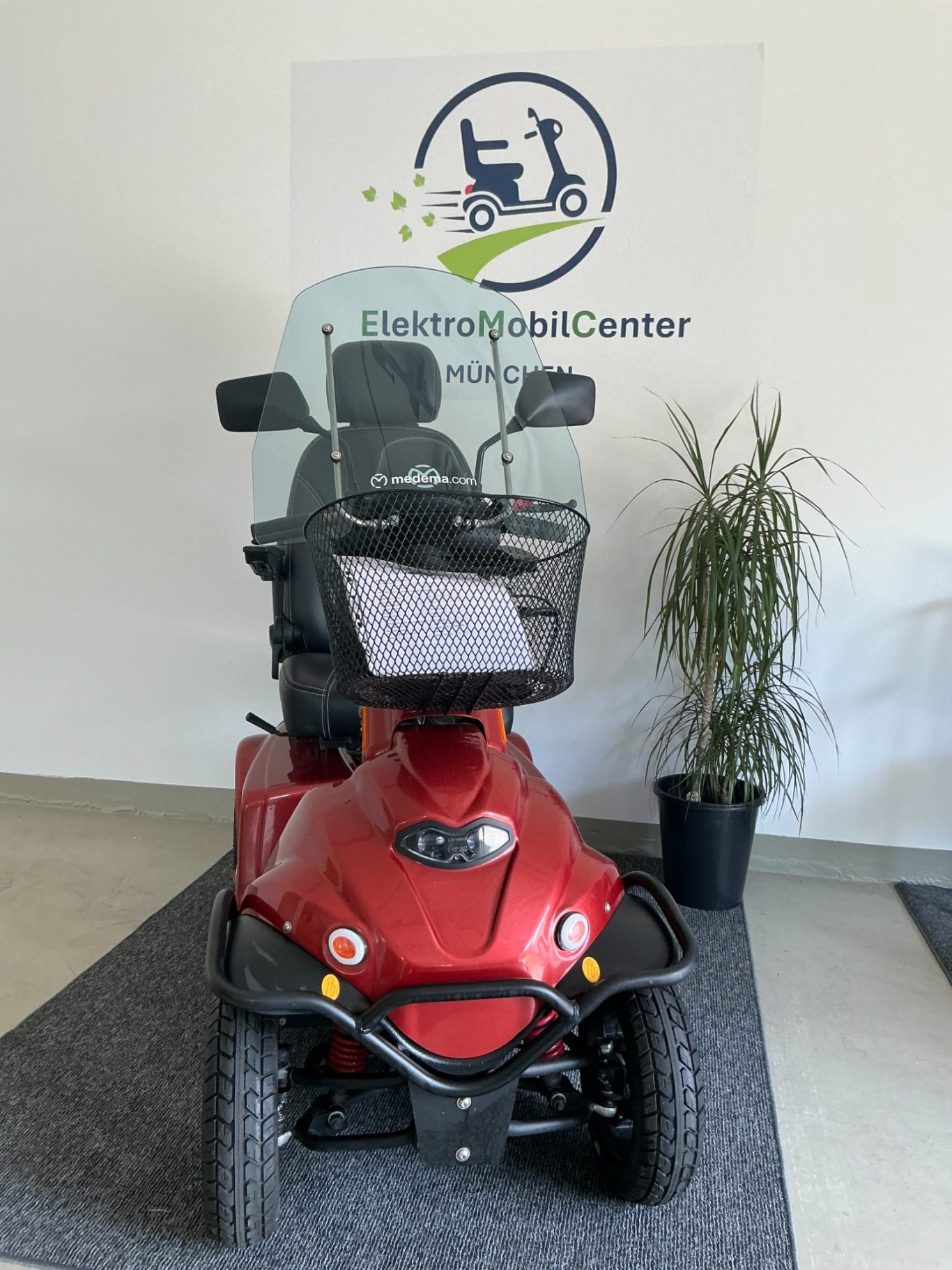 Roter Elektroroller mit Windschutzscheibe und Korb, vor einer Wand mit Logo.